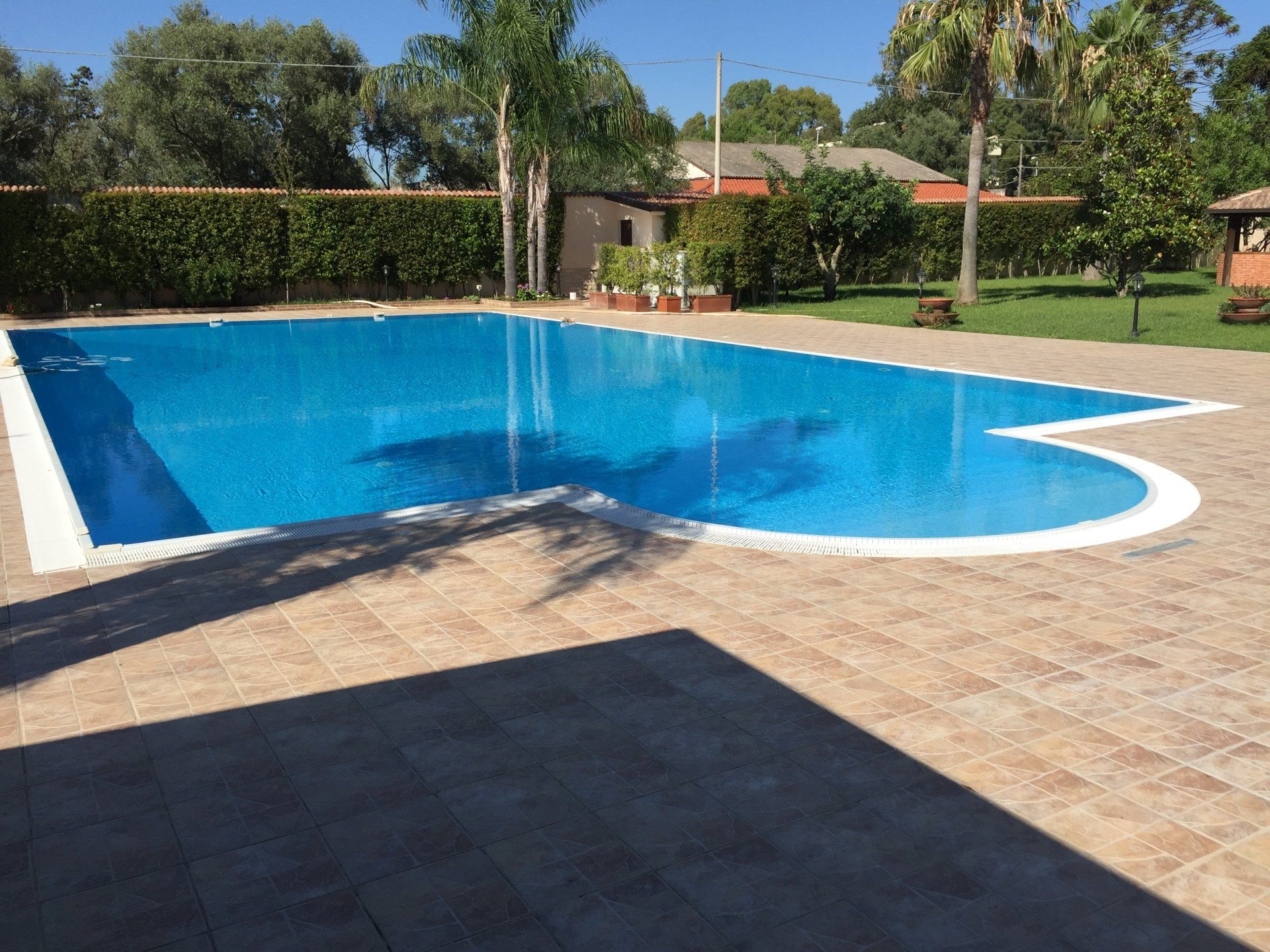 Piscina con acqua azzurra circondata da piastrelle beige e da un paesaggio verde.