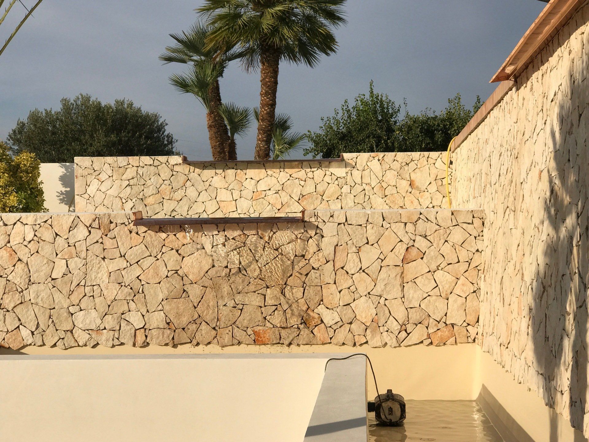Muro in pietra con cascata, palme e piscina.