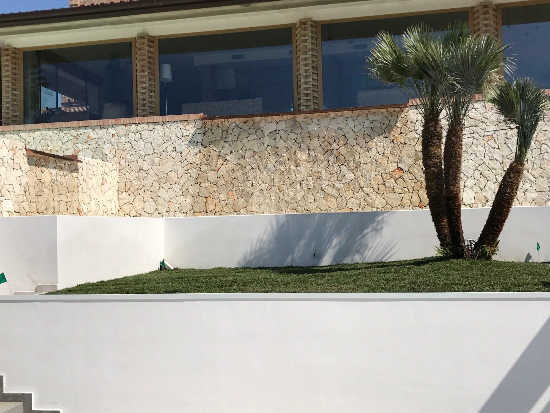 Muro di pietra con finestre nella parte superiore, palme e muro bianco di fronte.