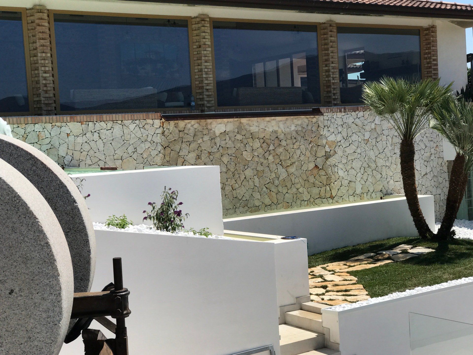 Esterno di un edificio con ampie finestre, pareti bianche e facciata in pietra. È visibile una palma.