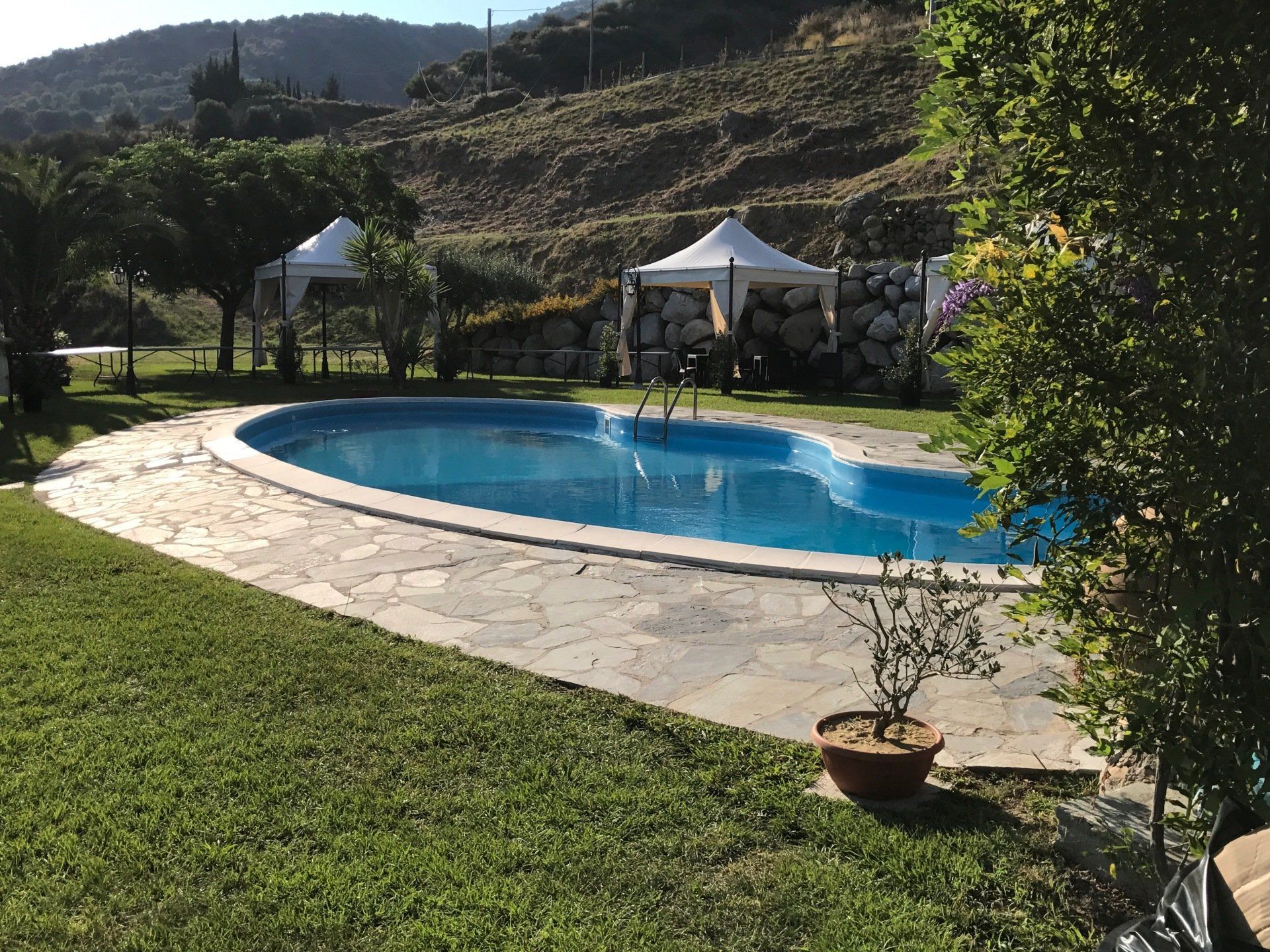 Piscina con patio in pietra, gazebo e prato erboso, con colline sullo sfondo.