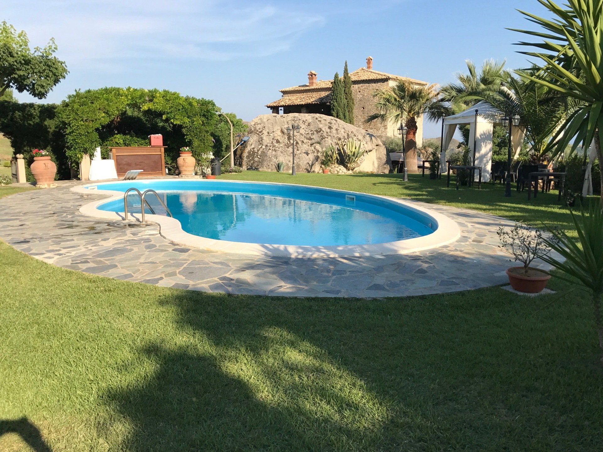 Piscina circondata da un prato verde, con un patio in pietra e un edificio sullo sfondo. Giornata di sole.