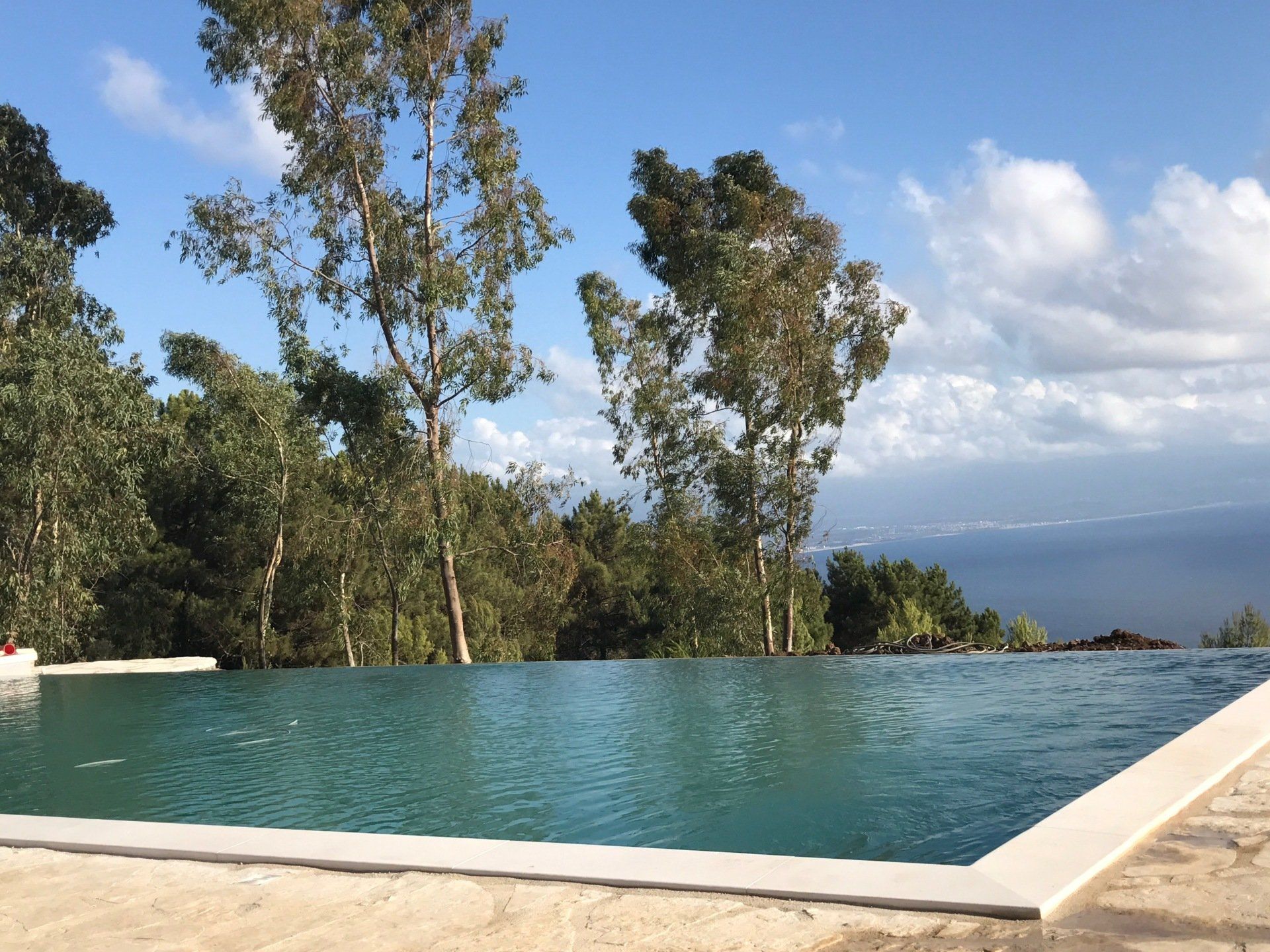 Piscina a sfioro con vista sugli alberi e sull'acqua sotto un cielo azzurro.