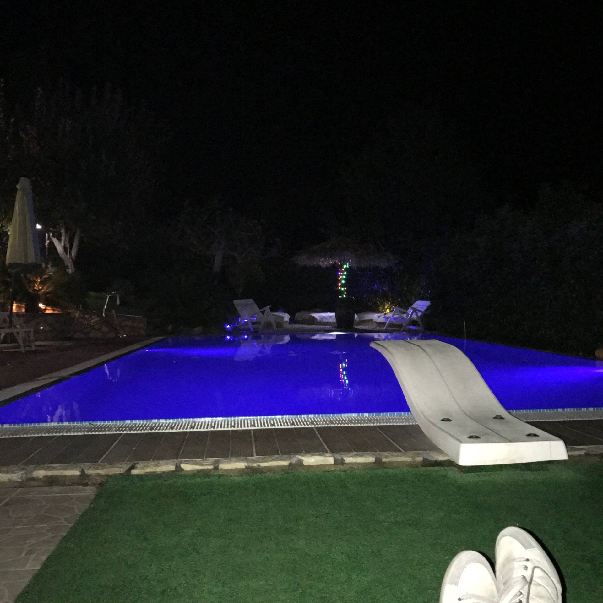 Piscina di notte con luci blu, lettini e uno scivolo. Erba verde in primo piano.