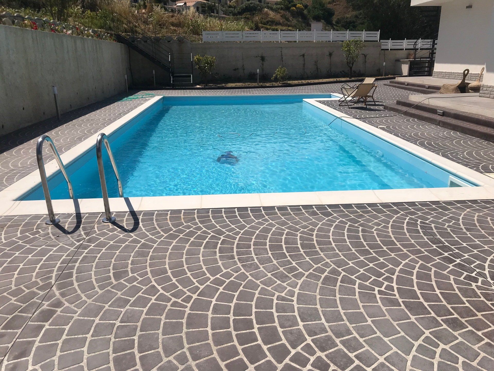 Piscina con acqua azzurra e patio in pietra, circondata da un muro e un po' di verde.