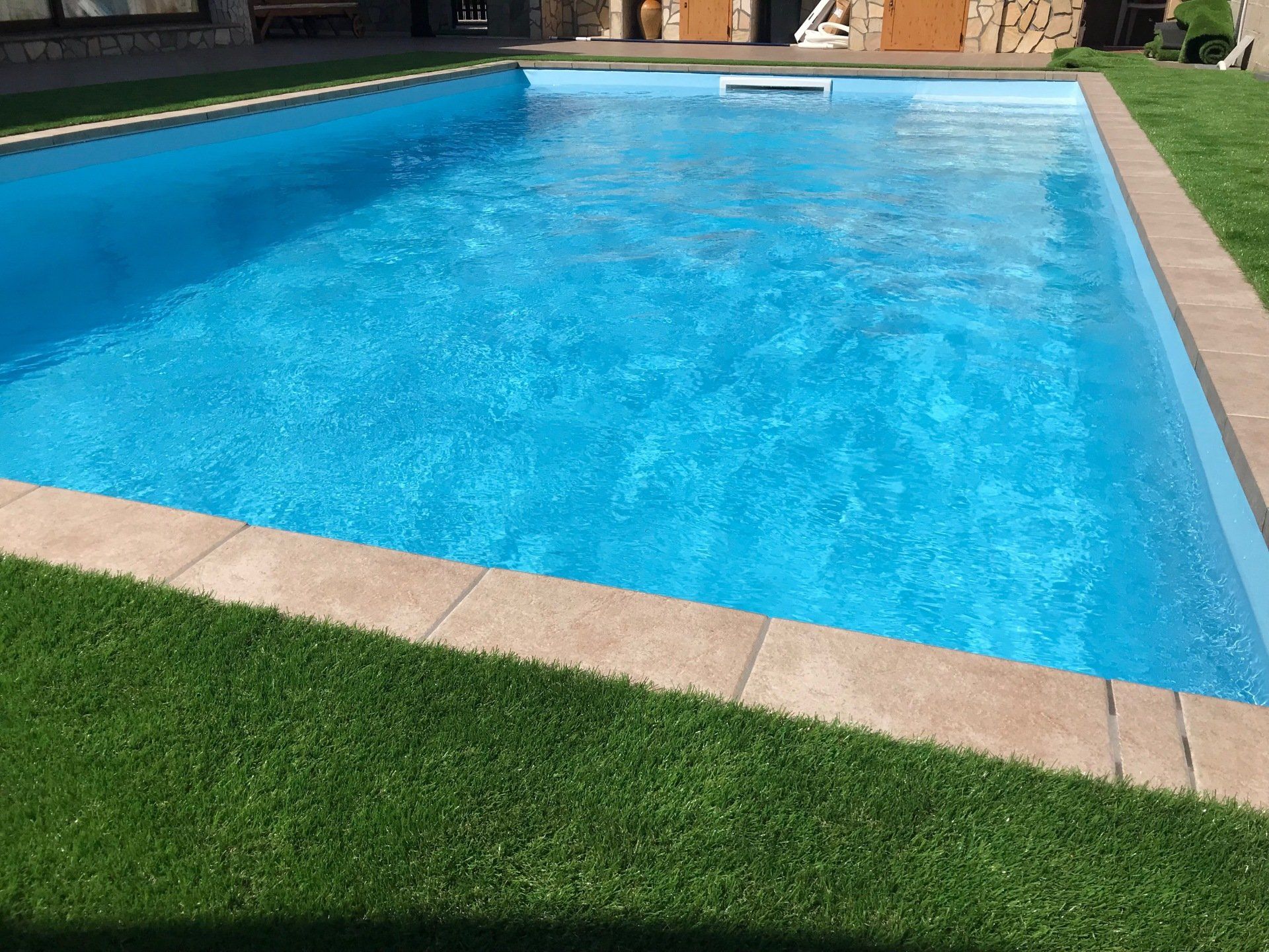 Piscina rettangolare con acqua azzurra accanto a prato verde e bordo in pietra marrone.