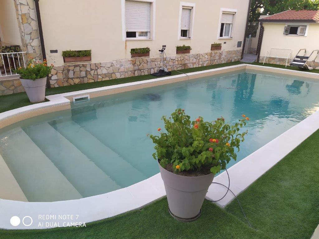 Piscina con gradini, piante in vaso e un edificio chiaro sullo sfondo. Il prato artificiale verde circonda la piscina.