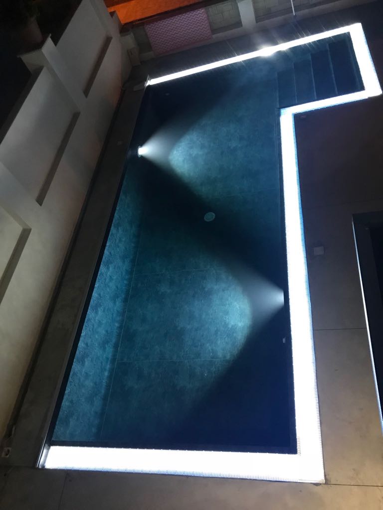 Una piscina rettangolare di notte, illuminata da faretti e luci di bordo, circondata da muri di edifici.