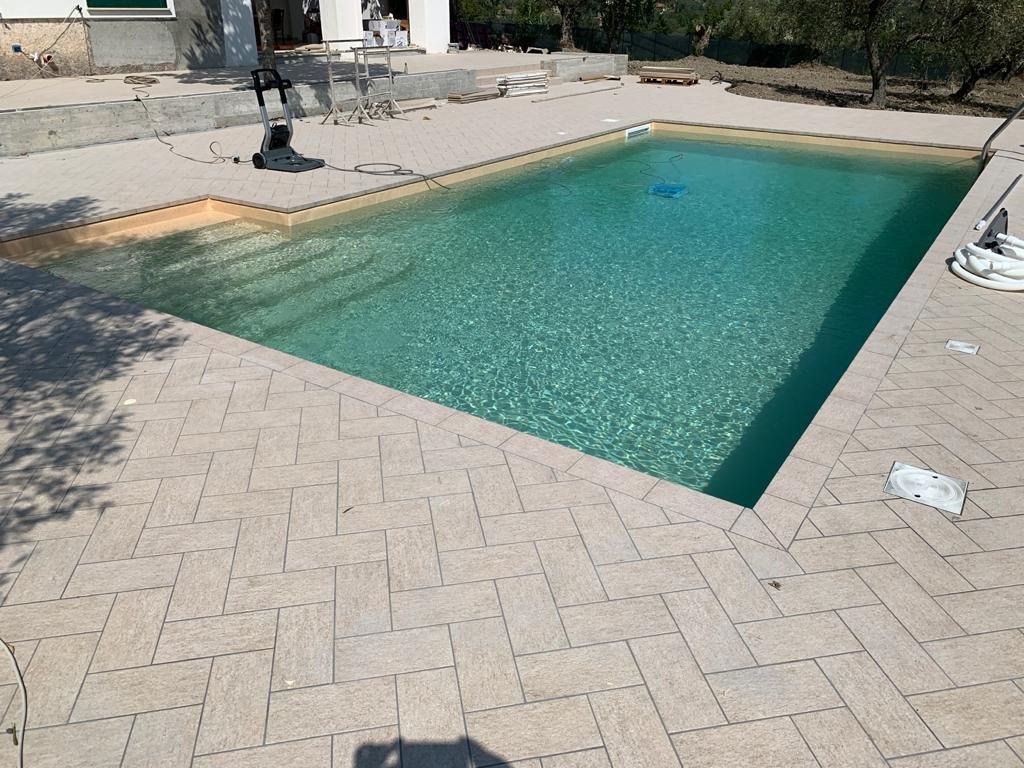 Piscina con acqua limpida, circondata da lastricati beige. In piscina è presente un robot pulitore.