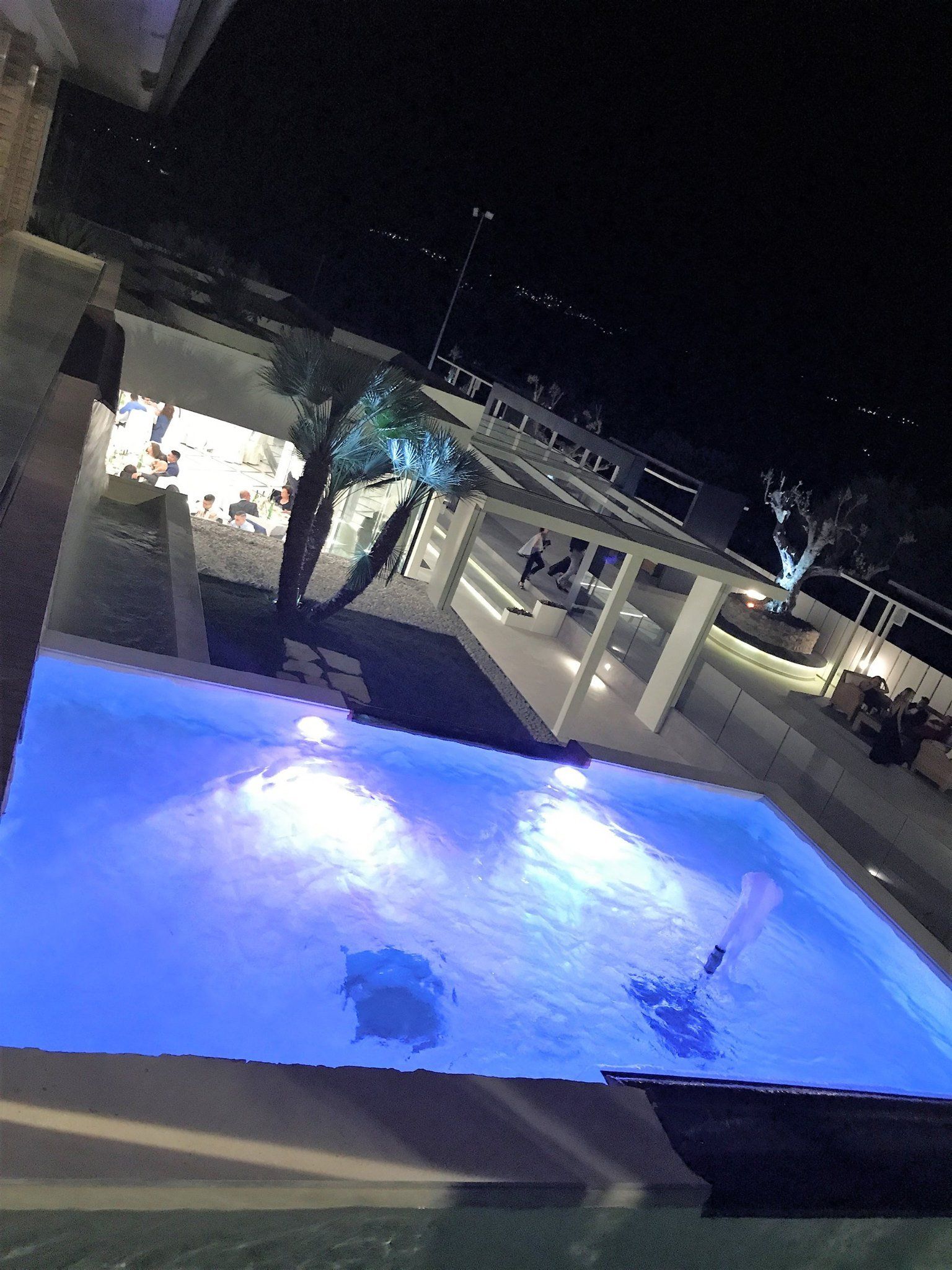 Vista notturna di una piscina rettangolare con luci blu, vicino a un pergolato bianco e ad alcuni alberi.