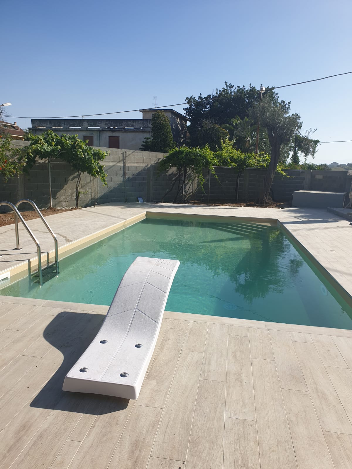 Piscina con trampolino, terrazza in cemento e recinzione circostante in una giornata di sole.