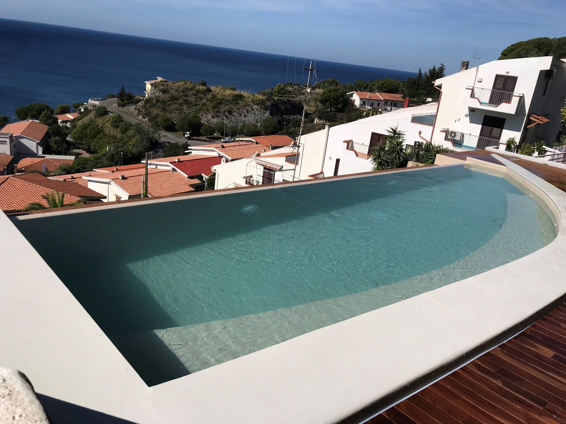 Una piscina a sfioro con vista sull'oceano, incorniciata dai tetti e da un paesaggio soleggiato.