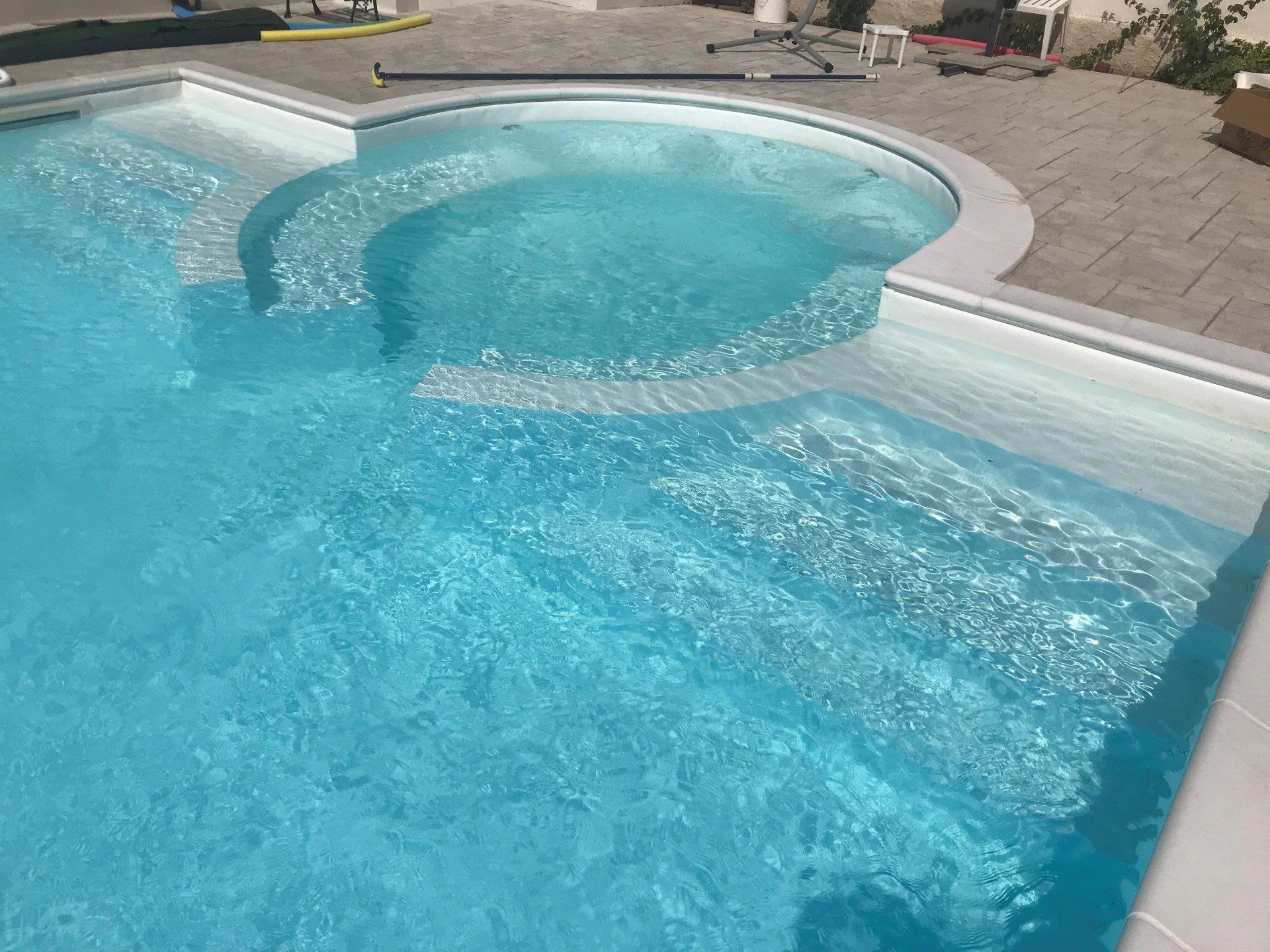 Piscina blu con gradini integrati e sezione curva. Circondata da pietra grigia e acqua.