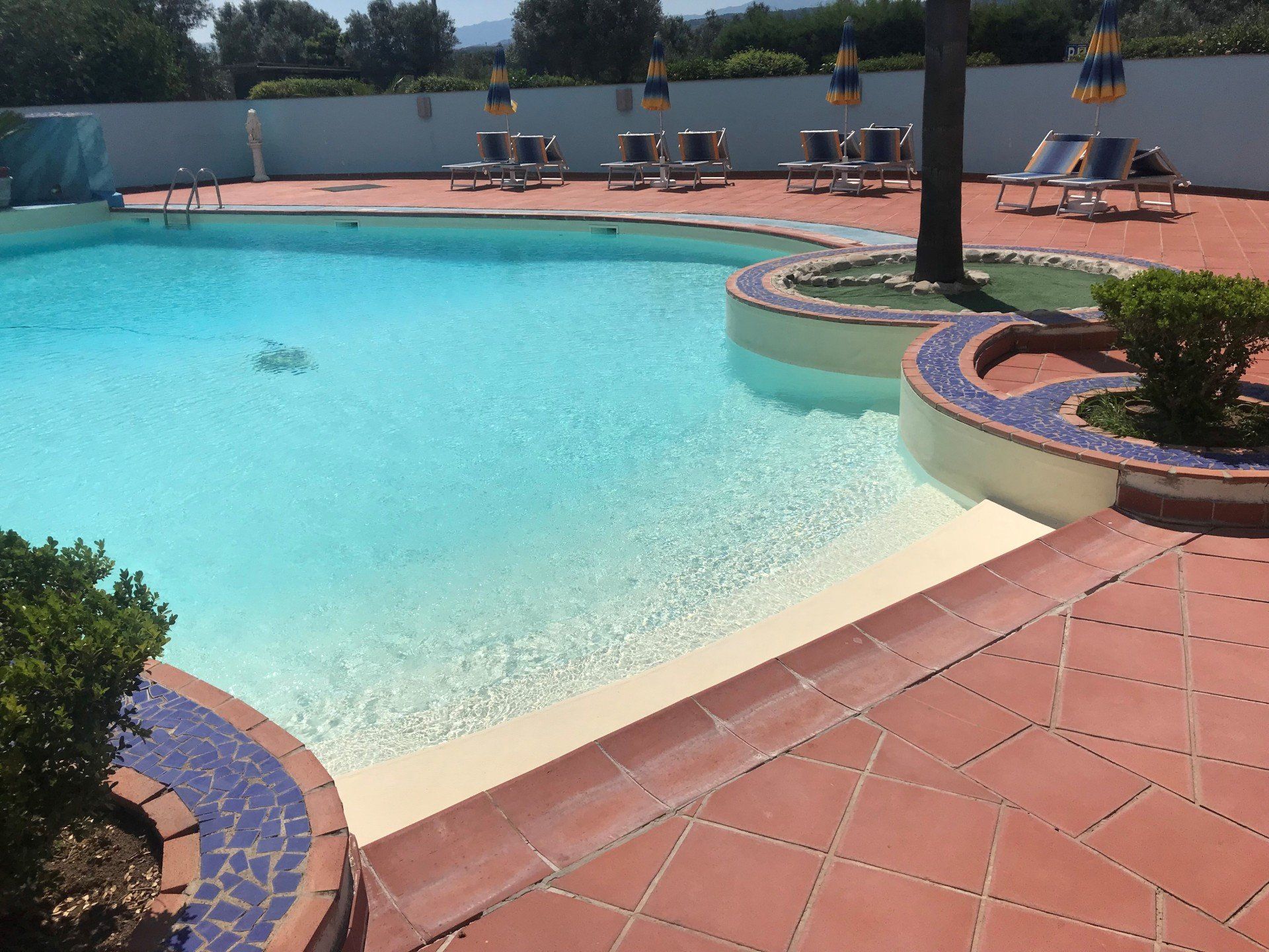 Piscina con acqua blu, patio con piastrelle rosse e sedie a sdraio.