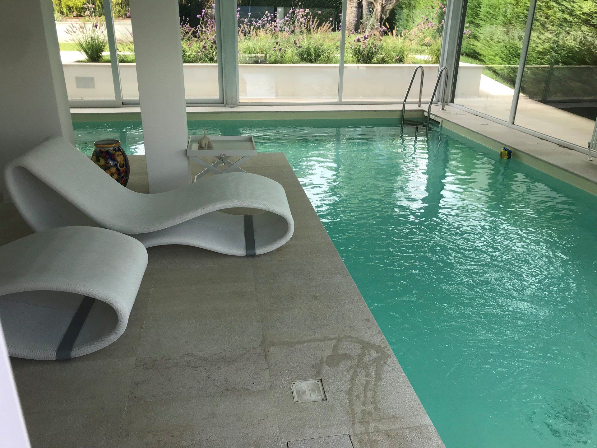 Una poltrona bianca e un tavolino sono posizionati accanto a una piscina, all'interno di uno spazio luminoso e chiuso.