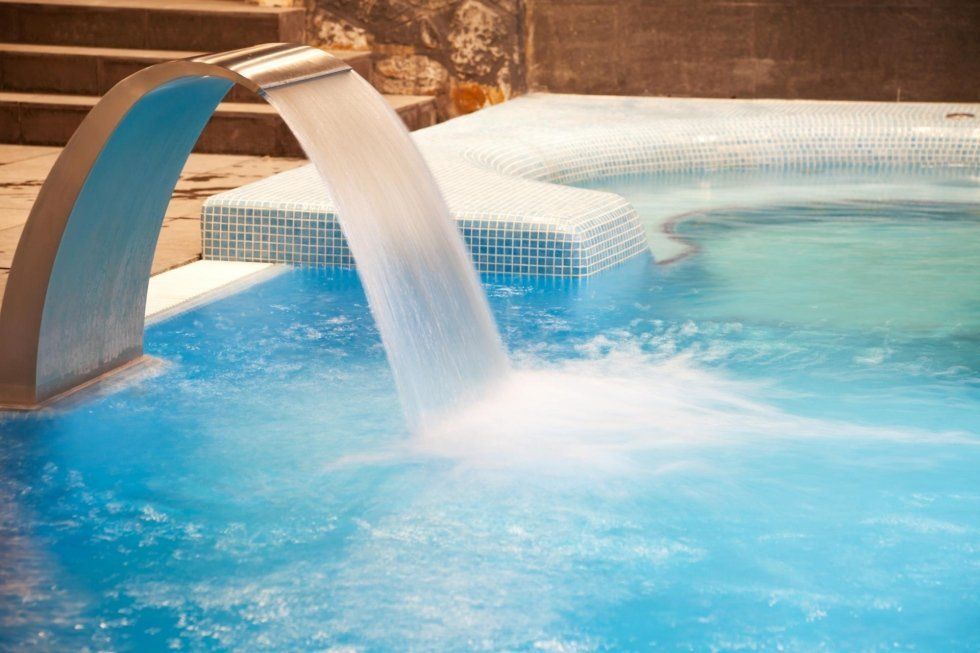 Piscina con cascata; acqua azzurra, piastrelle bianche, arco in acciaio inossidabile, gradini in pietra sullo sfondo.