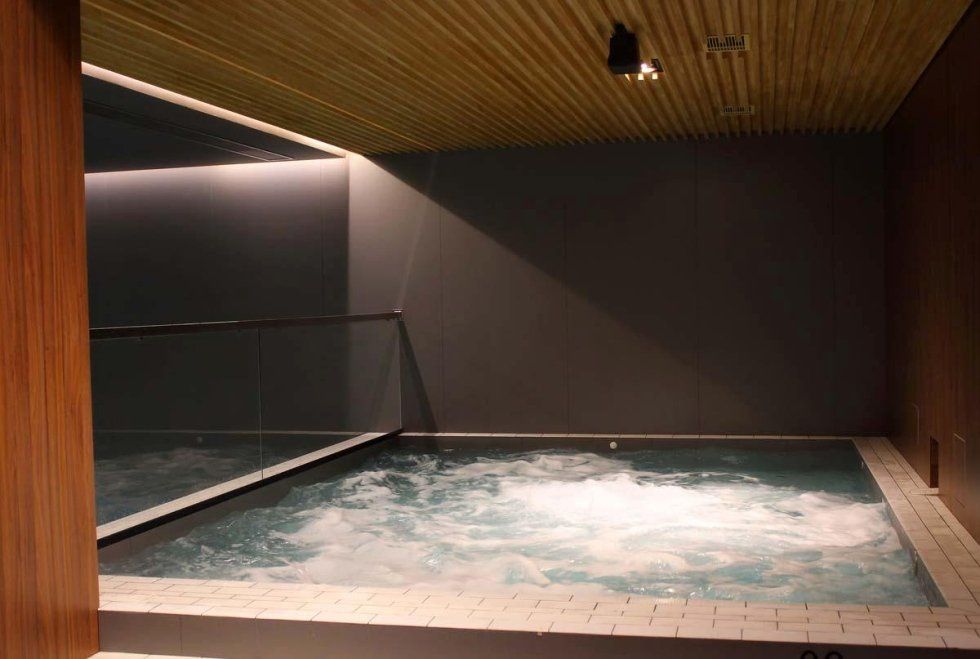 Jacuzzi interna con acqua gorgogliante, circondata da pareti grigio scuro, finiture in legno e soffitto a specchio.