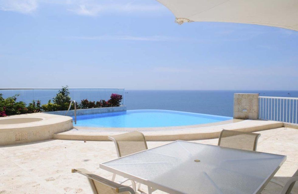 Piscina con vista sull'oceano; patio con tavolo, sedie e ombrellone sotto un cielo azzurro e soleggiato.