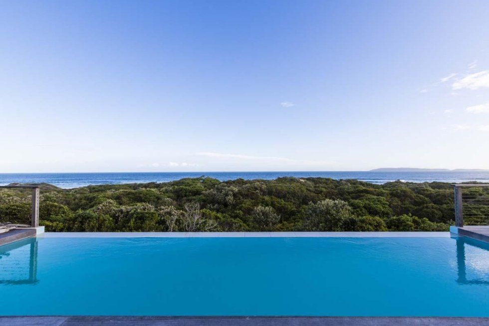 Piscina a sfioro con vista sull'oceano e sul fitto fogliame verde sotto un cielo azzurro e terso.