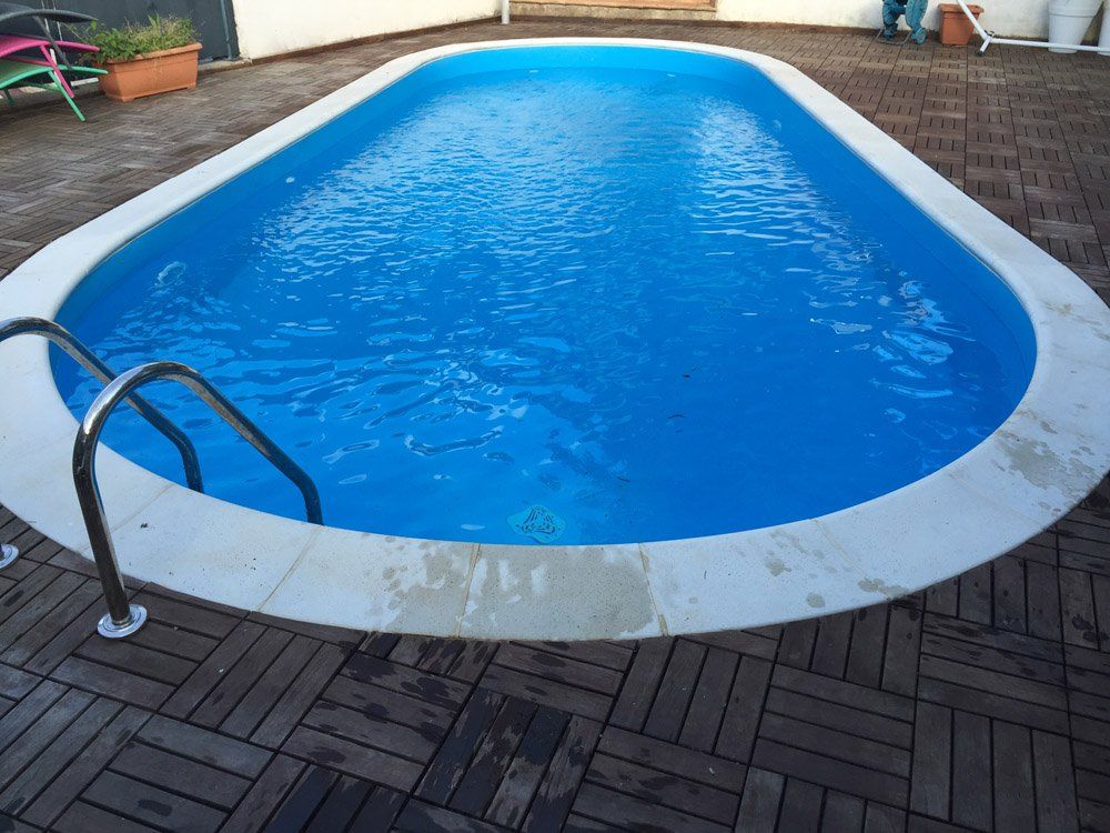 Piscina di forma ovale con acqua azzurra e bordo in cemento bianco, circondata da una terrazza in legno scuro.