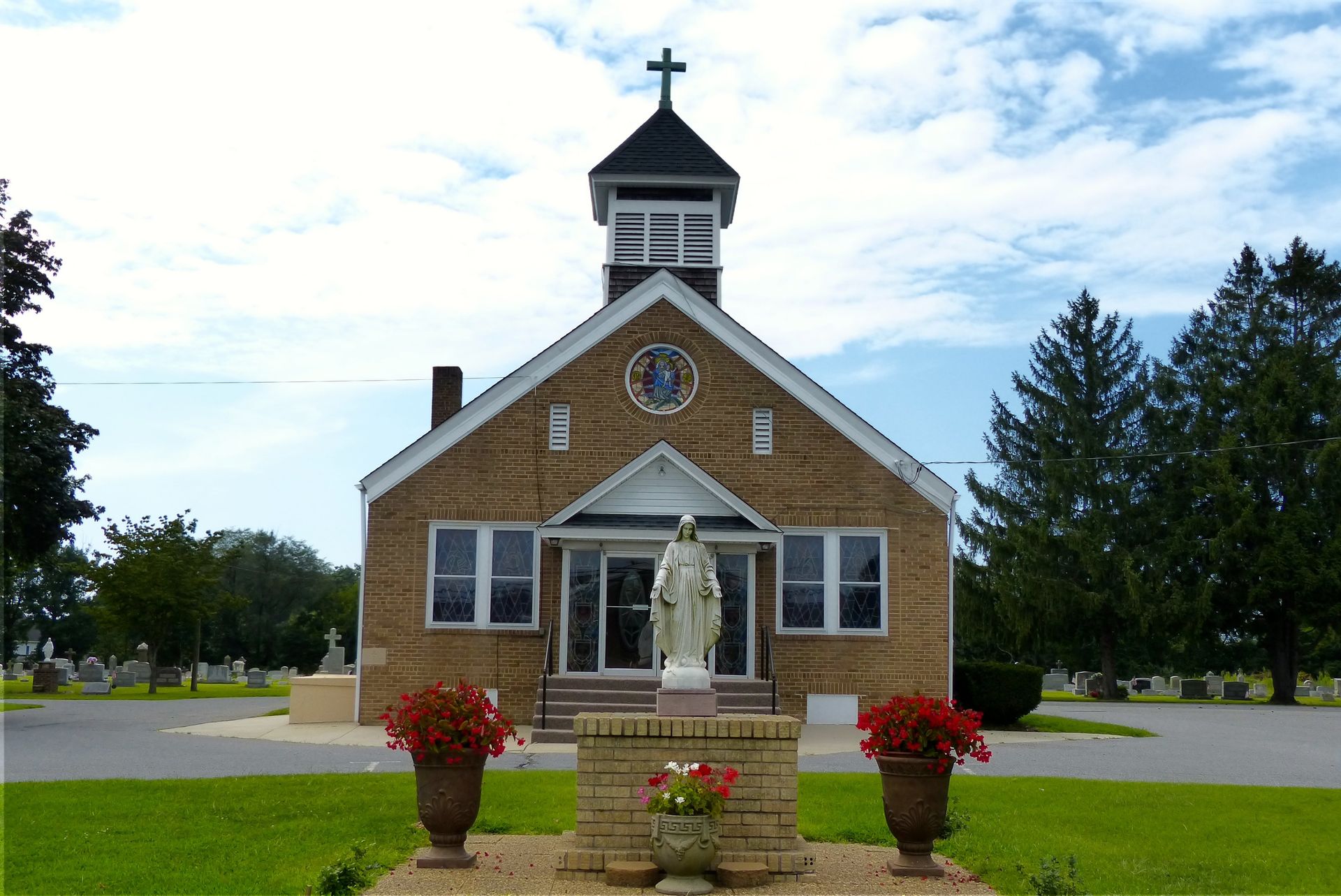 Stone chapel with statue of Virgin Mary, red flowers in urns, green grass, cloudy sky.