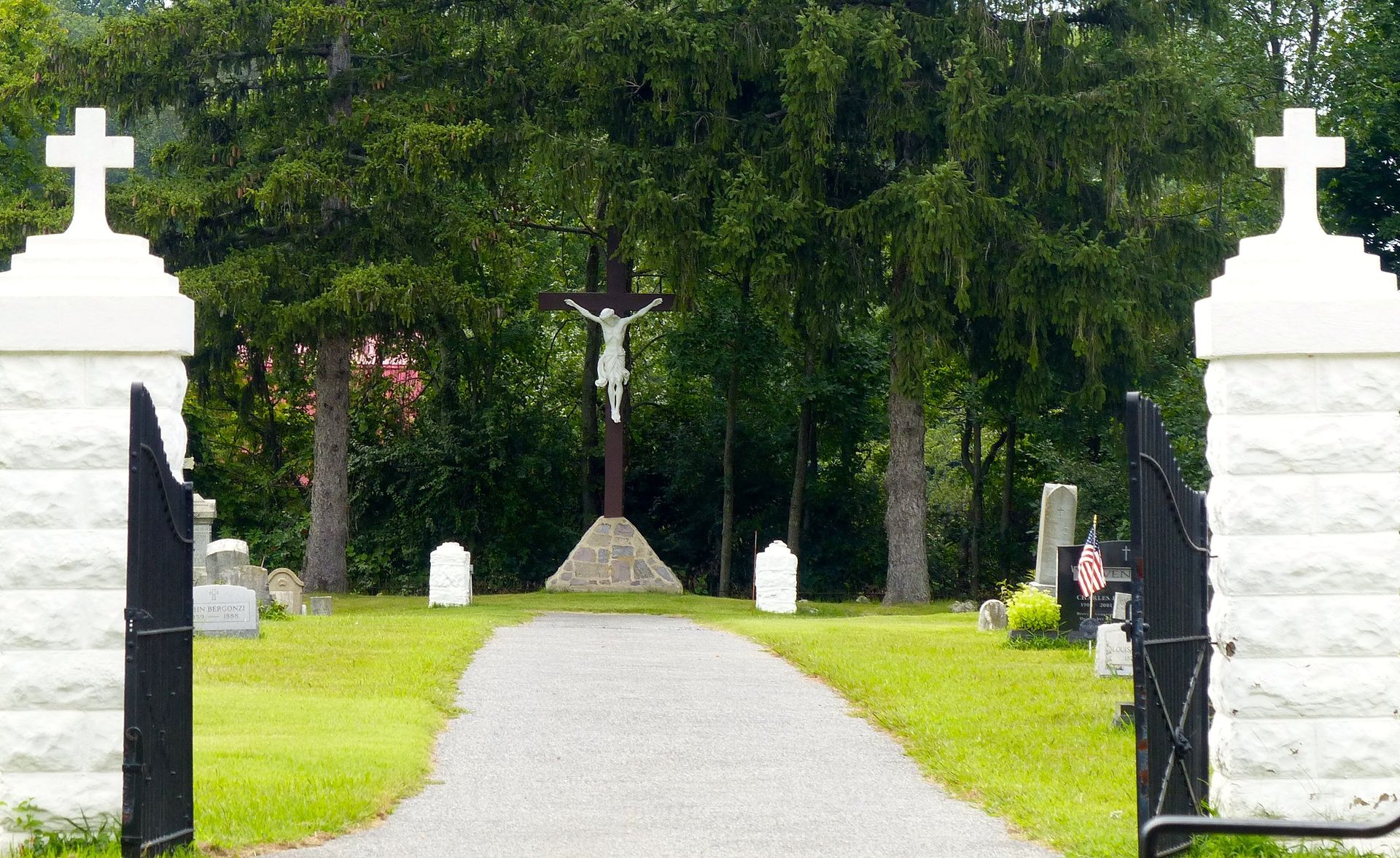 Entrance to a cemetery, white cross pillars framing a path leading to a crucifix in front of trees.