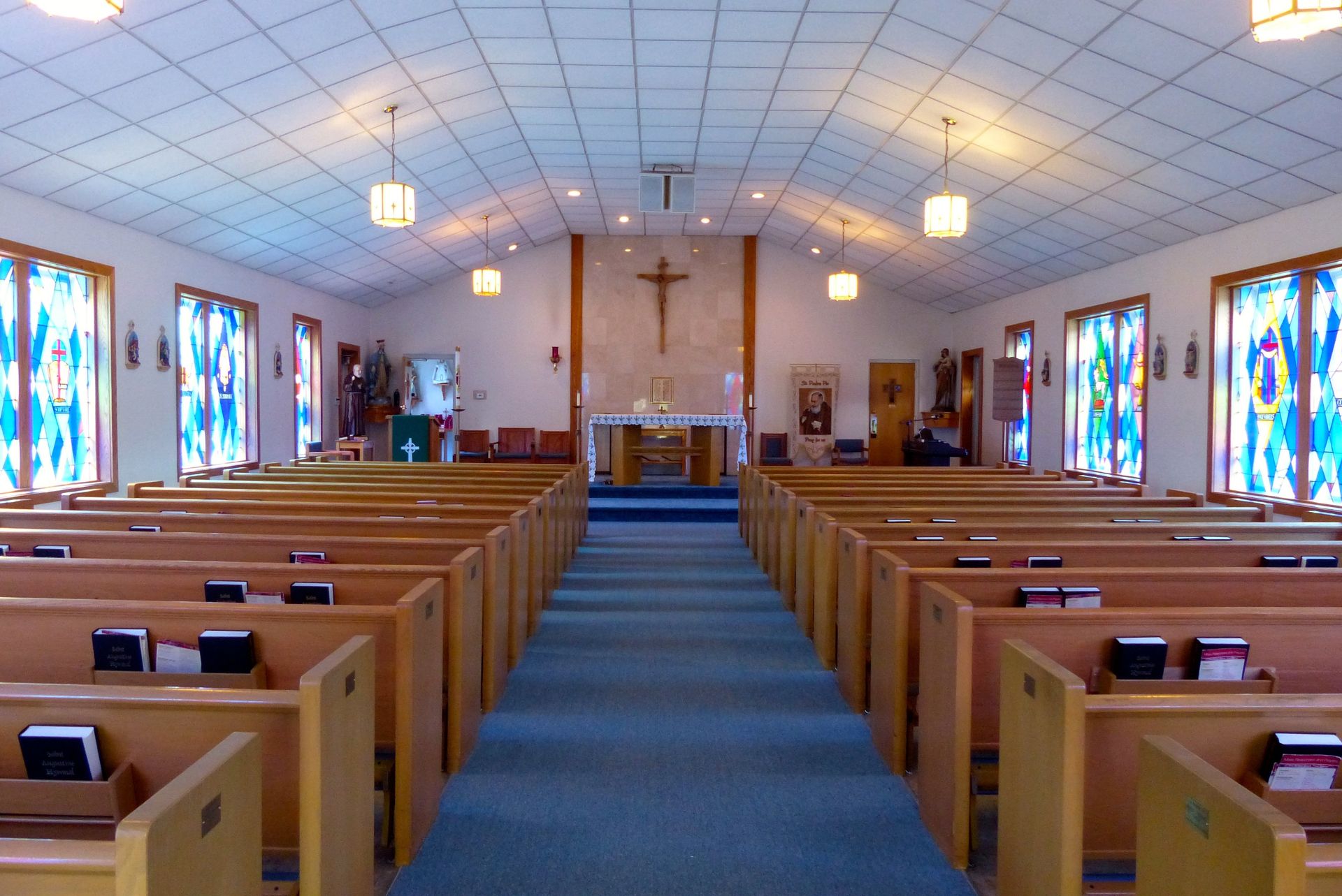 Interior of a church with rows of wooden pews, a blue carpeted aisle, and a crucifix on the altar.