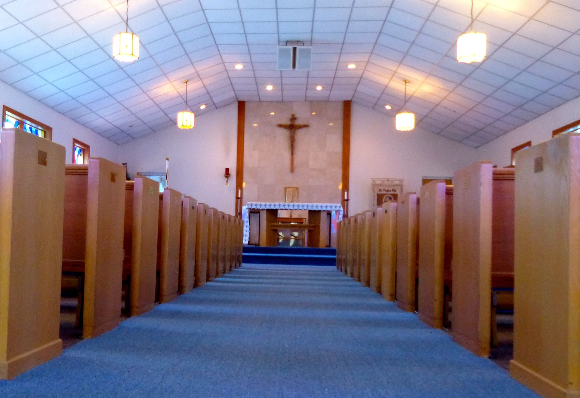 Interior view of a church with wooden pews, a blue carpeted aisle, and a crucifix above the altar.