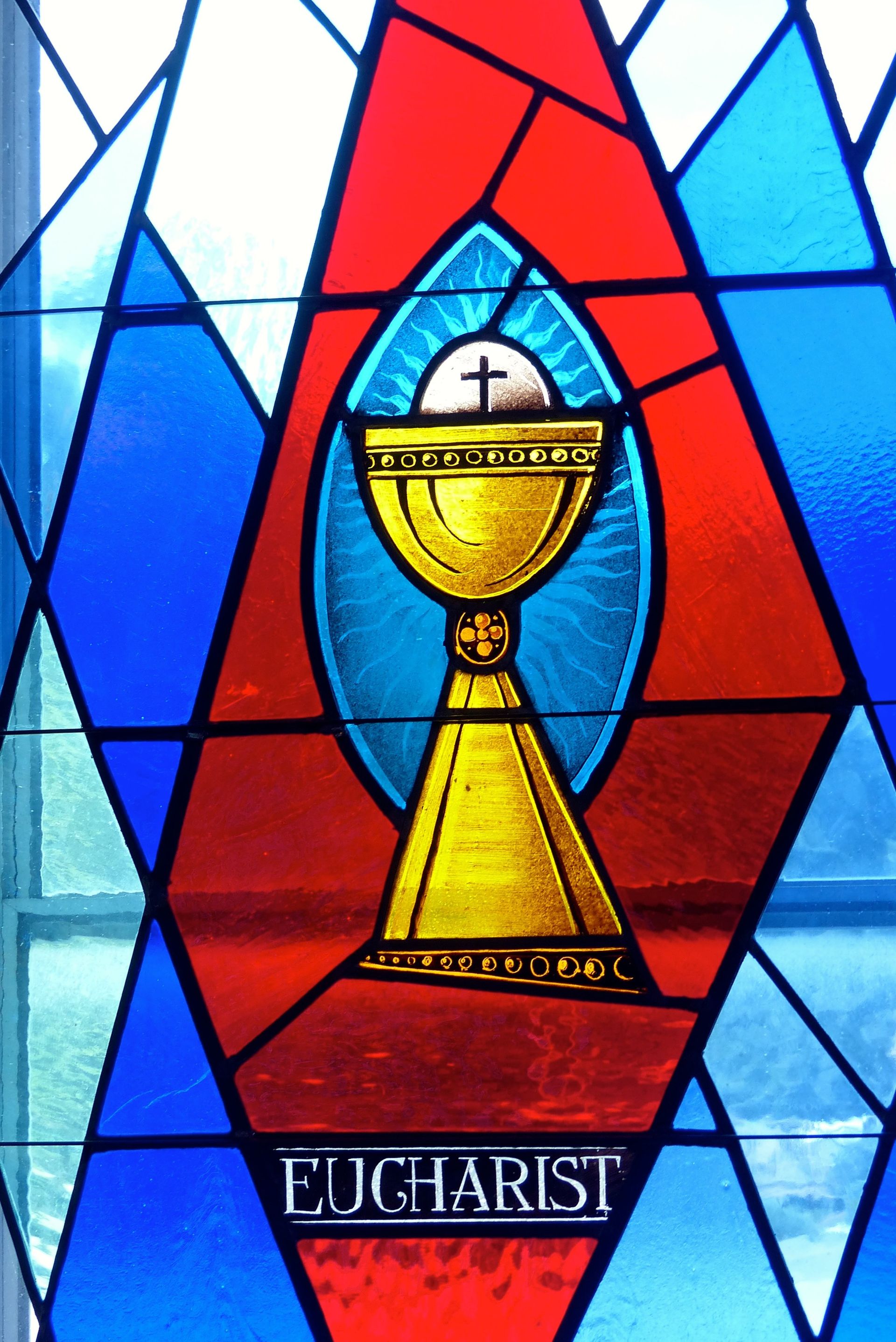 Stained glass window depicting a golden chalice with a host. Red and blue diamond-shaped background. Text reads 