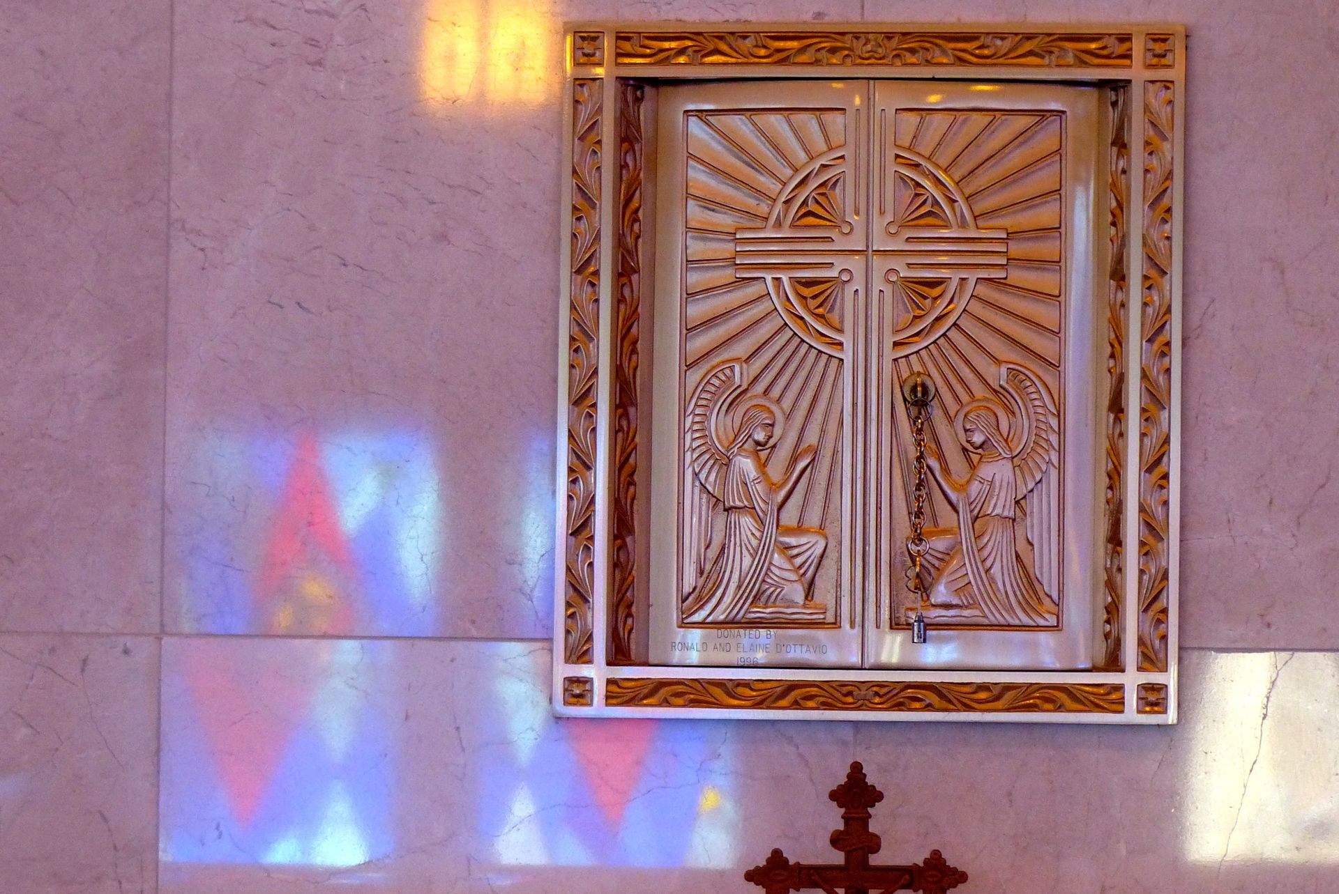 Ornate silver tabernacle on a wall with a cross design, stained-glass reflections.