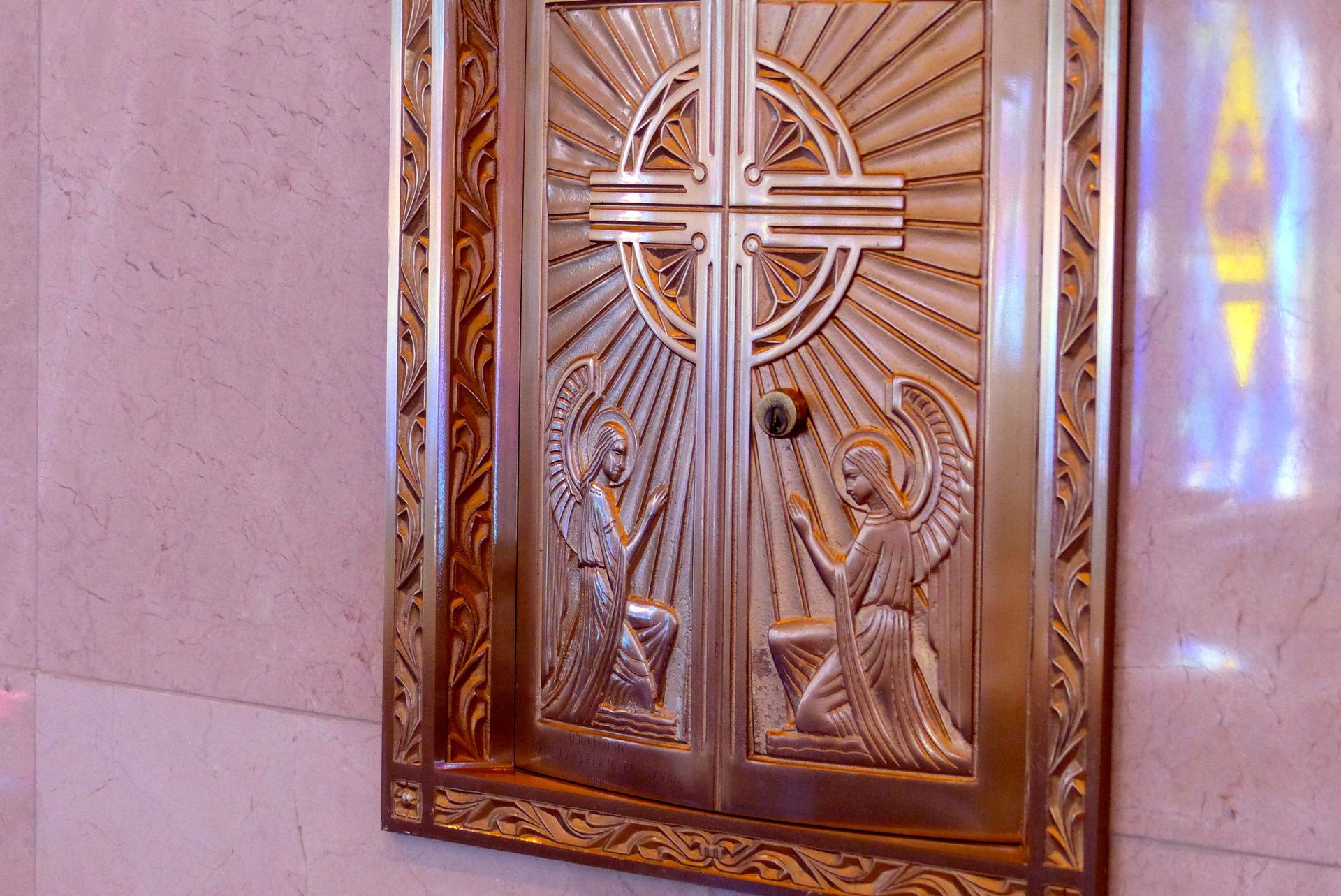 Gold-colored ornate doors with a cross design, flanked by angels, set in a light pink stone wall.