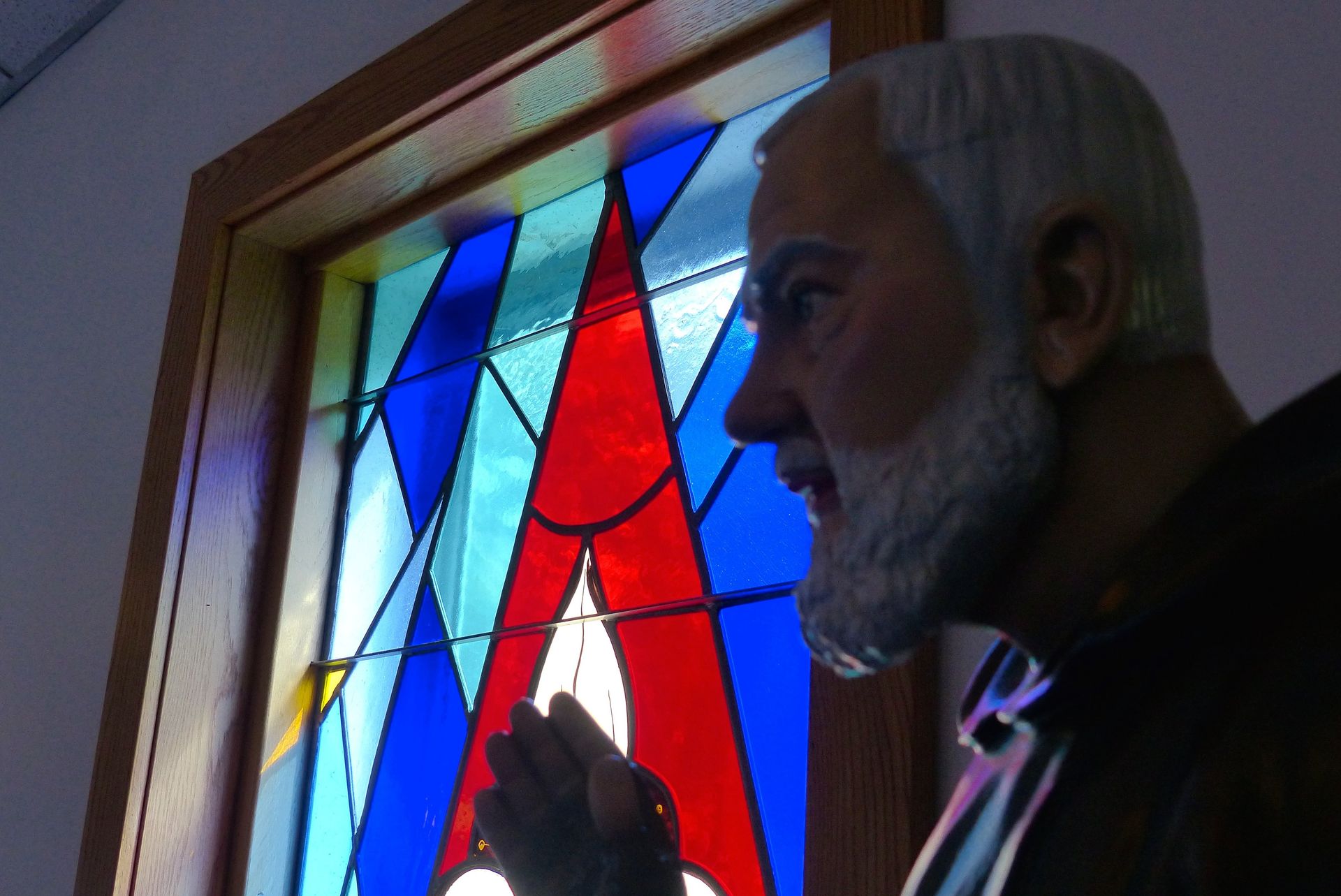 Statue of a man with white beard and hooded cloak, near a stained glass window with red, blue, and yellow panes.