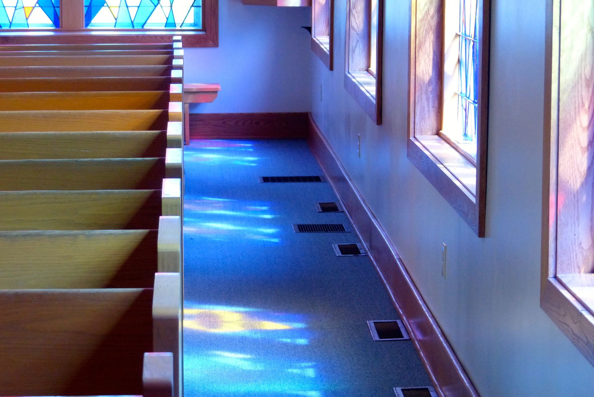 Church interior with wooden pews, stained glass windows, and a blue, textured floor reflecting light.