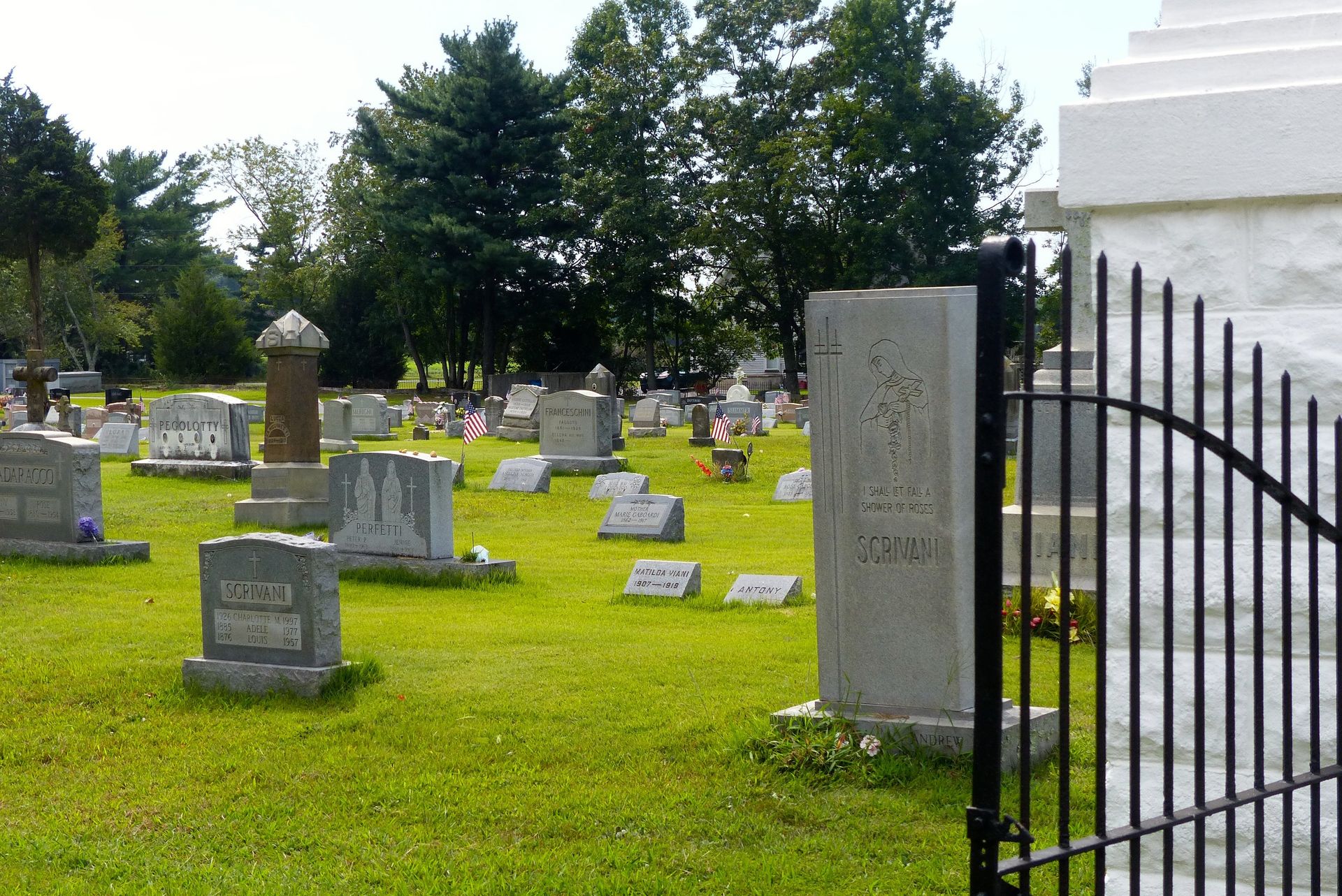 A cemetery with many headstones on green grass, trees in the background, gate in the foreground.