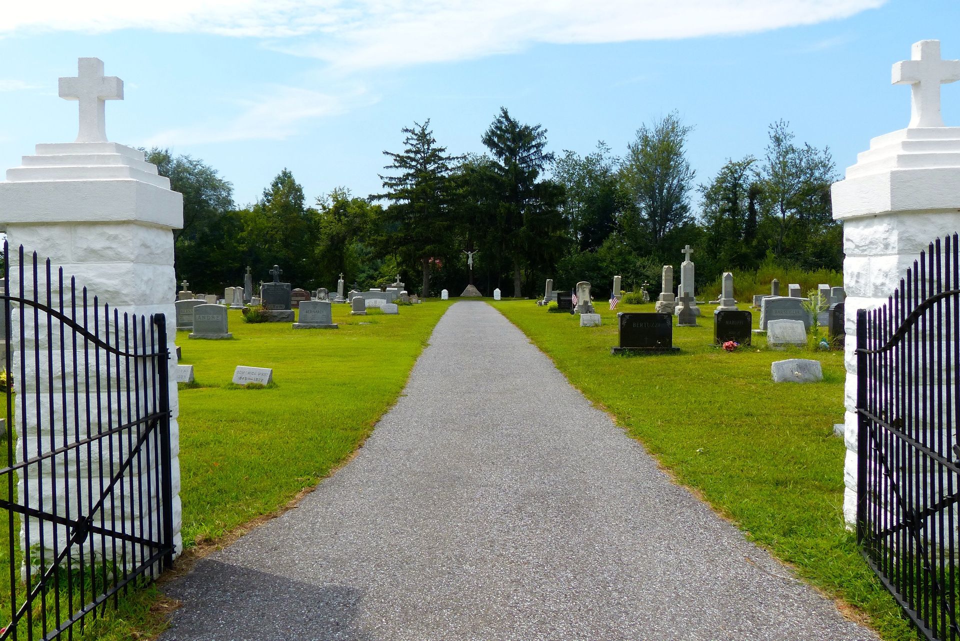 Gravel path leading into a cemetery, flanked by headstones on green grass. White gates and crosses. Blue sky.