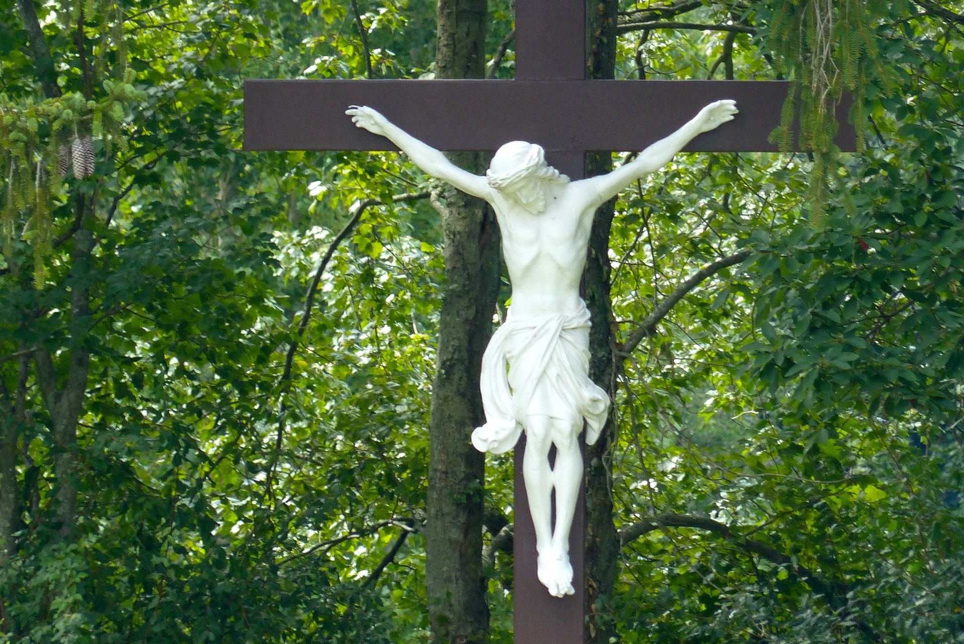 White statue of Jesus on a dark brown cross against a background of green trees.