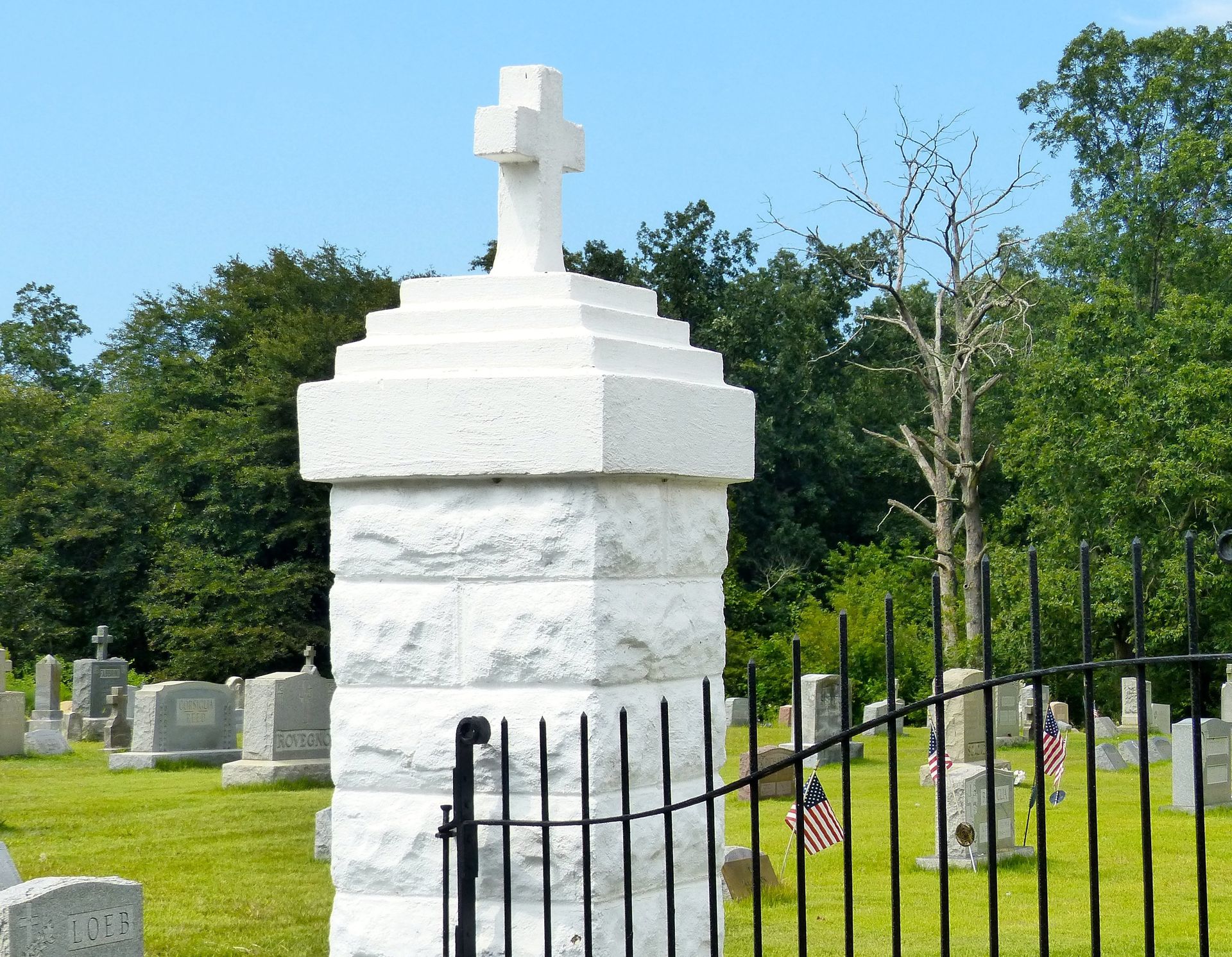 White stone pillar topped with a cross at a cemetery gate, green grass and headstones in the background.