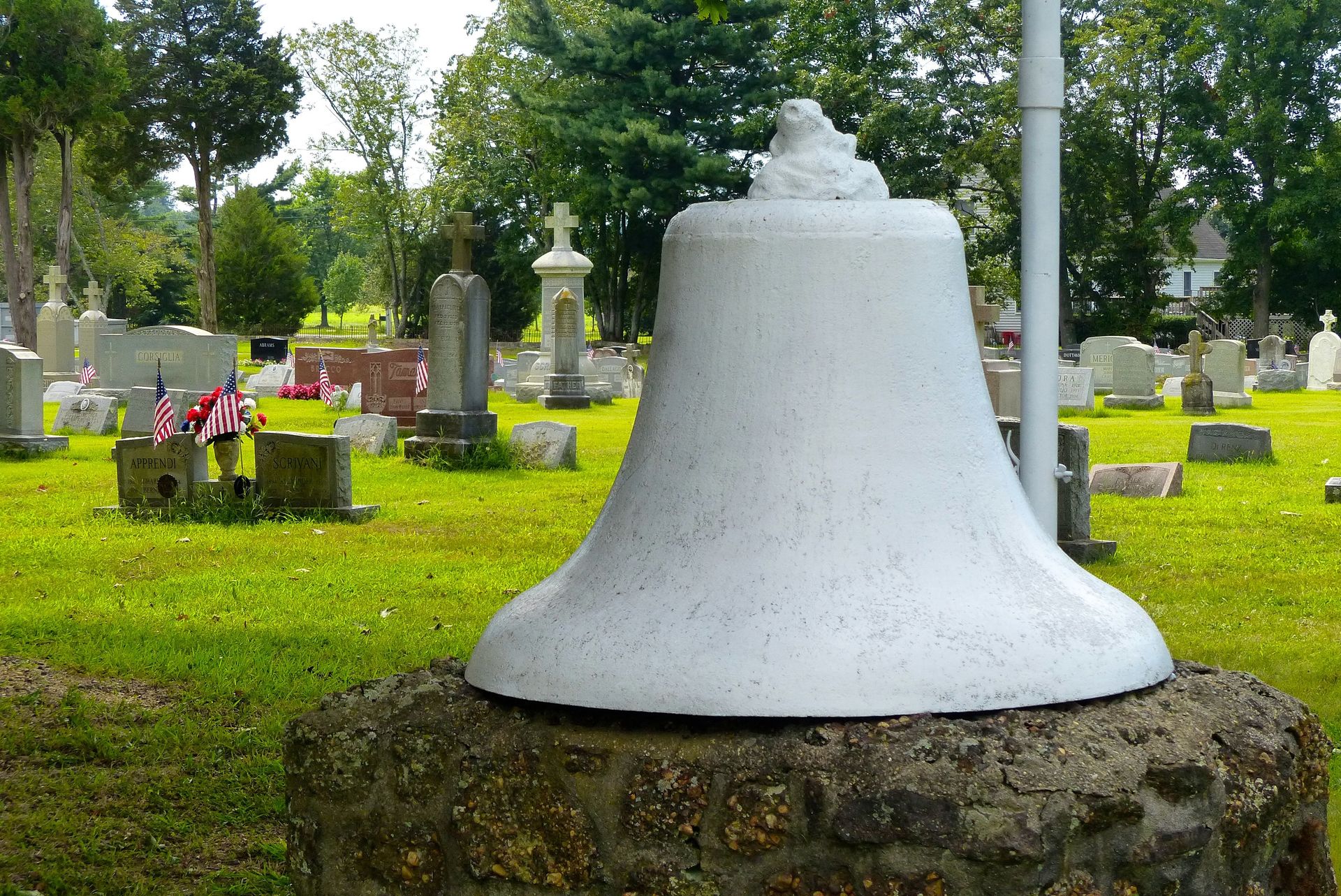 White bell on a stone base in a cemetery with headstones and flags; green grass and trees.
