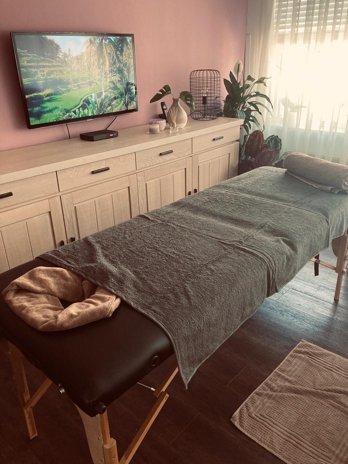 A massage table with a grey cover sits in a room with a light wood sideboard, a wall-mounted TV, and indoor plants.