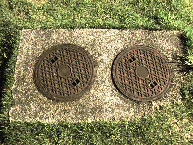 Two circular manhole covers set in a rectangular stone block, surrounded by green grass.