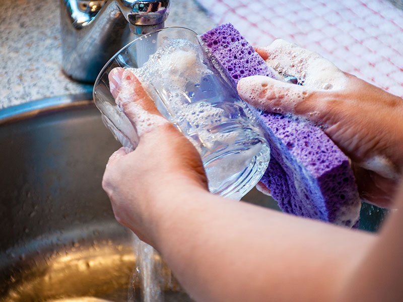 Hands washing a clear glass in a sink with a purple sponge.