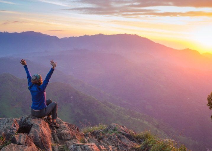 A woman sitting on a mountain top during sunrise, raising her arms up