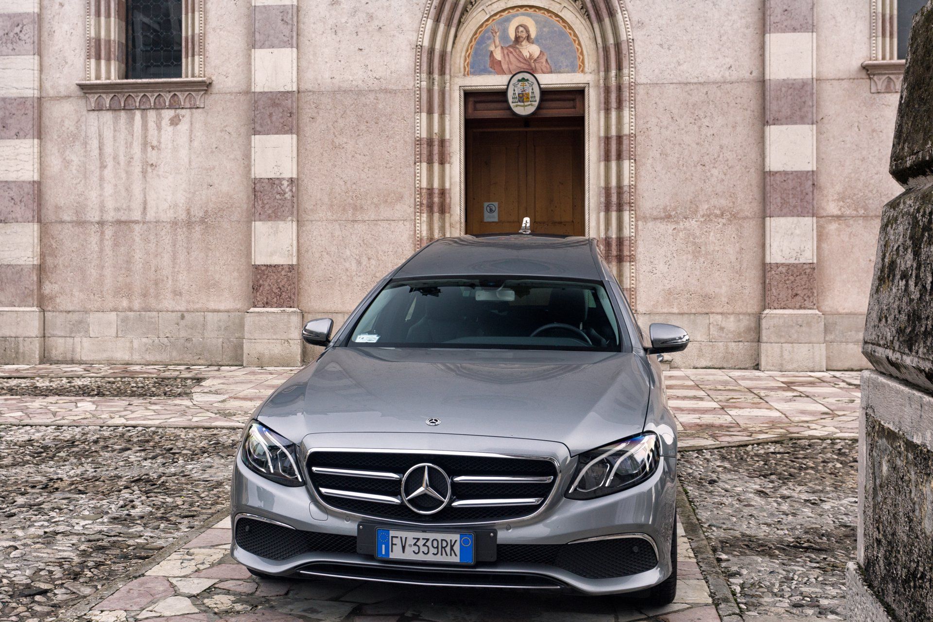 Carro funebre Mercedes color argento parcheggiato davanti a una chiesa, targa italiana.