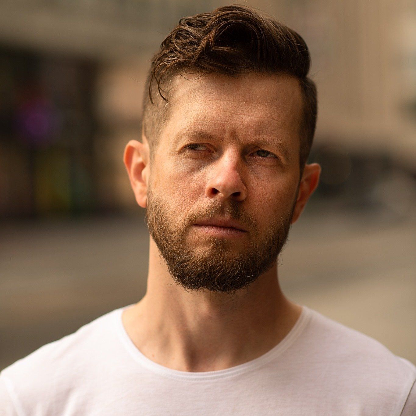 A man with a beard is wearing a white t-shirt and looking at the camera.