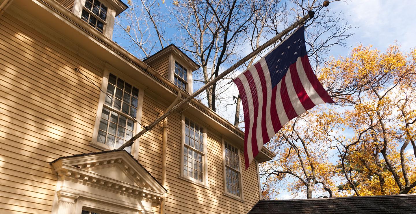 American flag hanging from a brick building beside autumn trees and blue sky