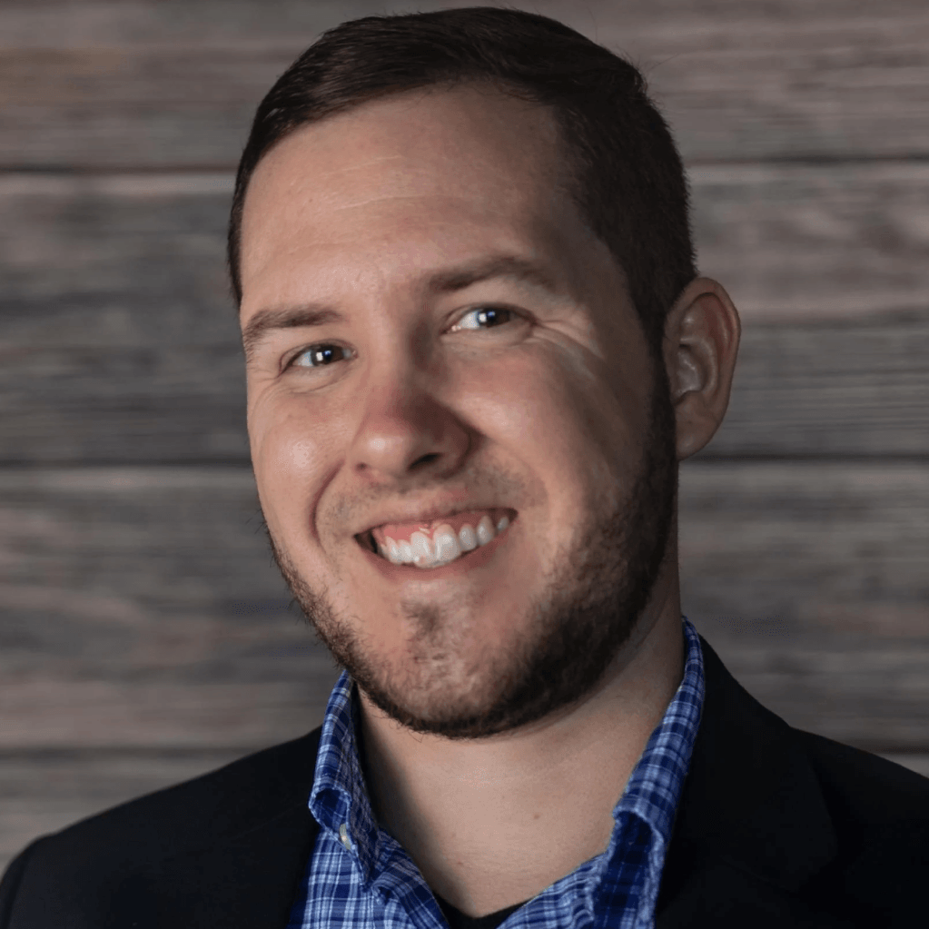 A man with a beard is smiling in front of a wooden wall.