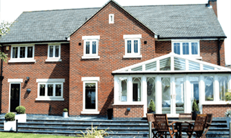 Large brick house with gray roof and white conservatory, wooden deck, and patio furniture outdoors