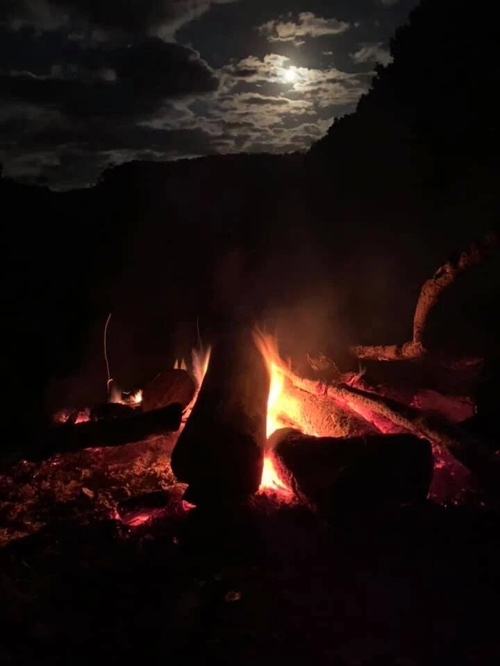 A person is sitting around a campfire at night with a full moon in the background.