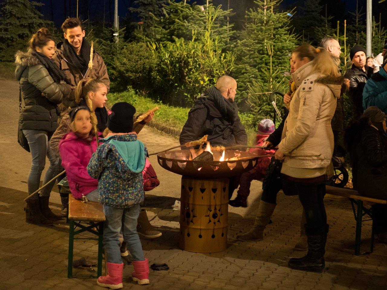 Mehrere Menschen sitzen oder stehen um ein Lagerfeuer und rösten Stockbrot