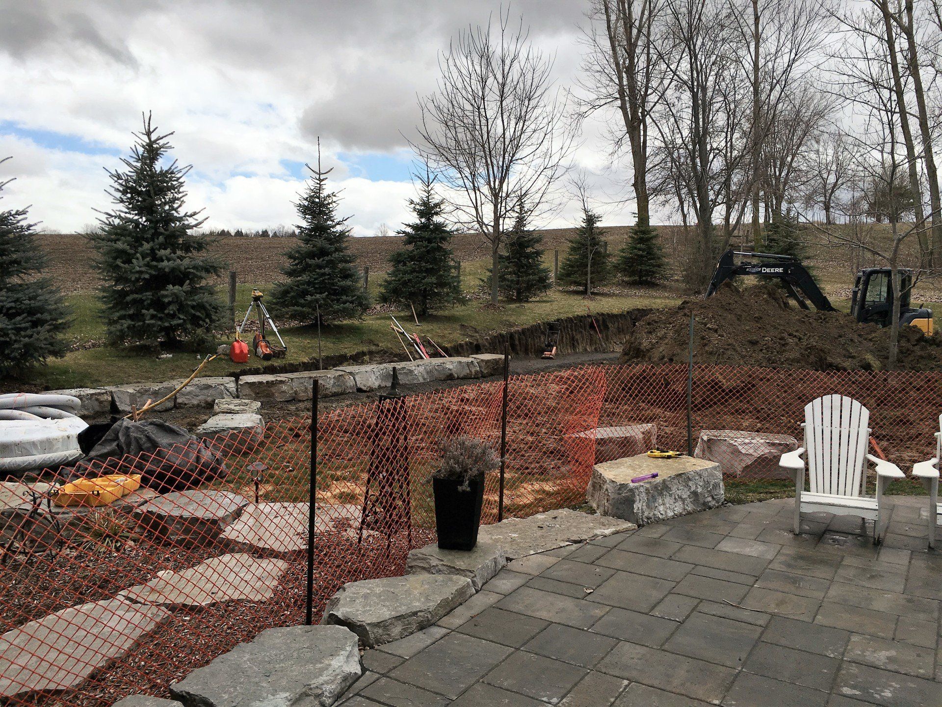 Construction site: Excavator digging near patio with safety fencing, trees, and sky.